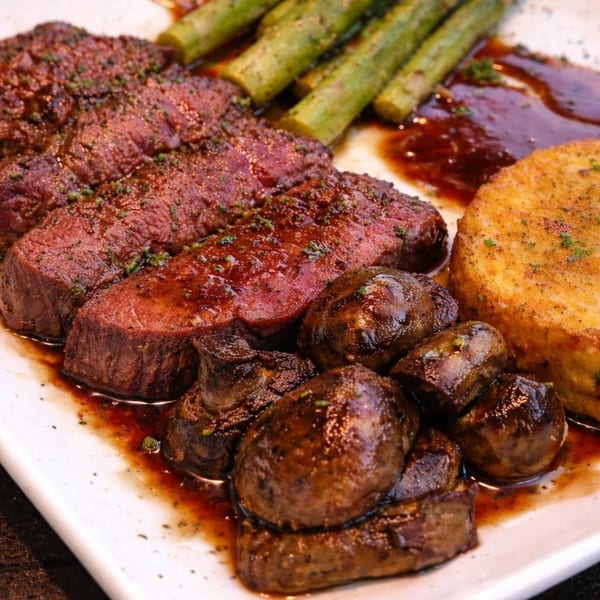 Sliced steak with asparagus, mushrooms, and a golden-brown side dish, drizzled with sauce.