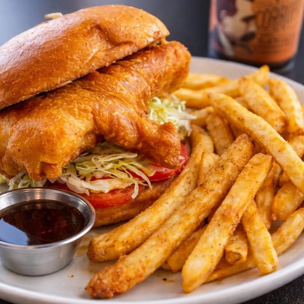 Crispy fried fish sandwich with lettuce, tomato, and a side of golden french fries and dipping sauce.