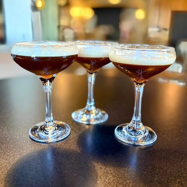 Three coupe glasses filled with dark cocktails topped with white foam on a dark table.