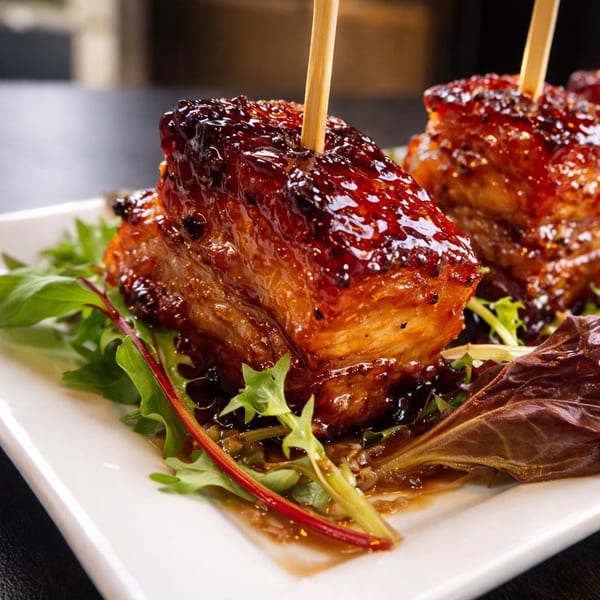 Two glazed pork belly bites with toothpicks, served on a bed of mixed greens.