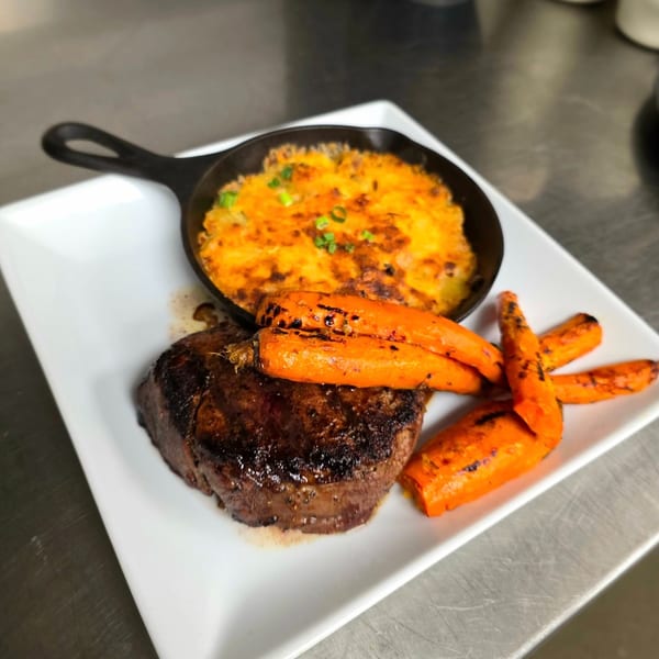 Seared steak with roasted carrots and a skillet of cheesy potatoes, garnished with chives.