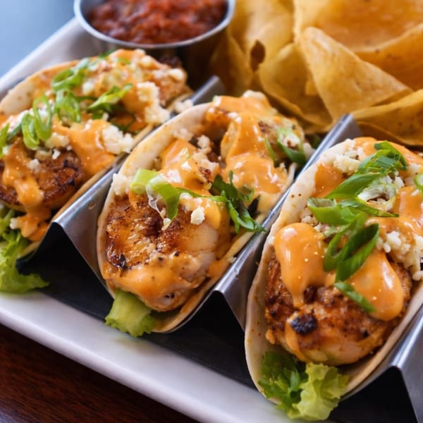 Three shrimp tacos with sauce, cheese, and green onions, served with chips and salsa.